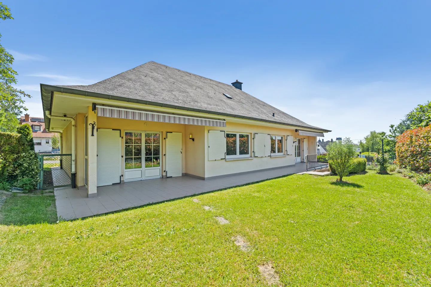 A yellow house with a gray roof, a covered patio, and white shutters, set against a bright blue sky and green lawn.