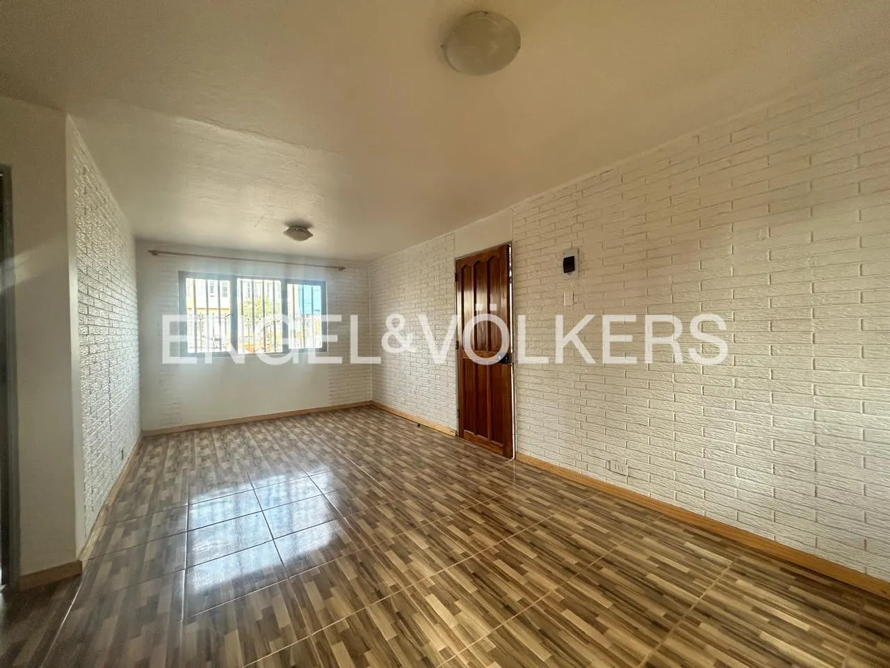 Empty room with wood-look tile floor, white brick walls, and a wood door. A window lets in natural light.