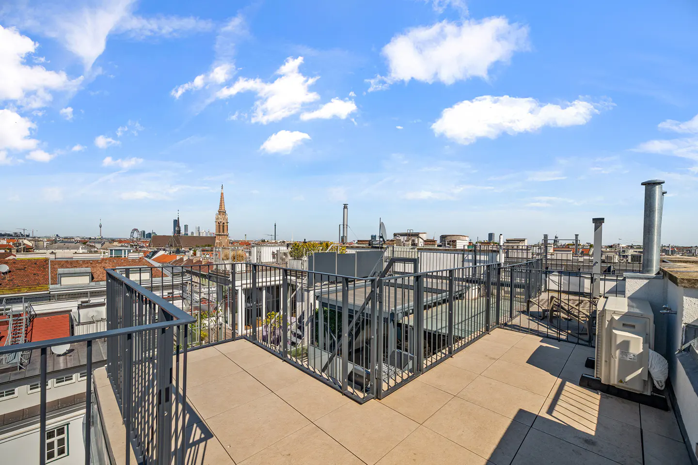 Rooftop terrace with gray metal railings and beige tiles, overlooking a city skyline under a blue sky with scattered clouds.