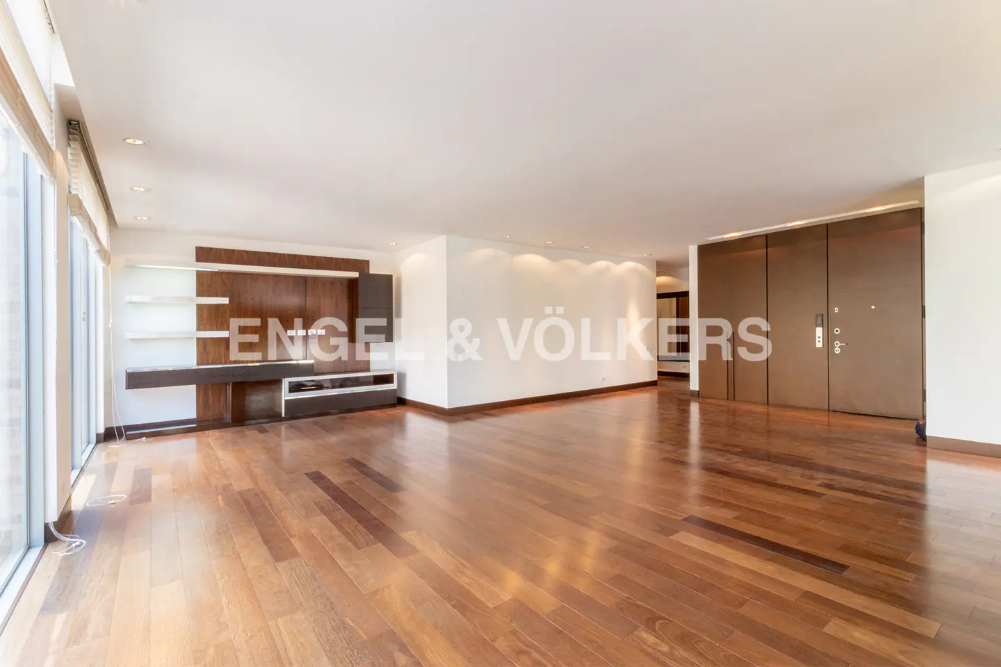 Bright, empty living room with hardwood floors, white walls, and a dark wood entertainment center. Large windows on the left.