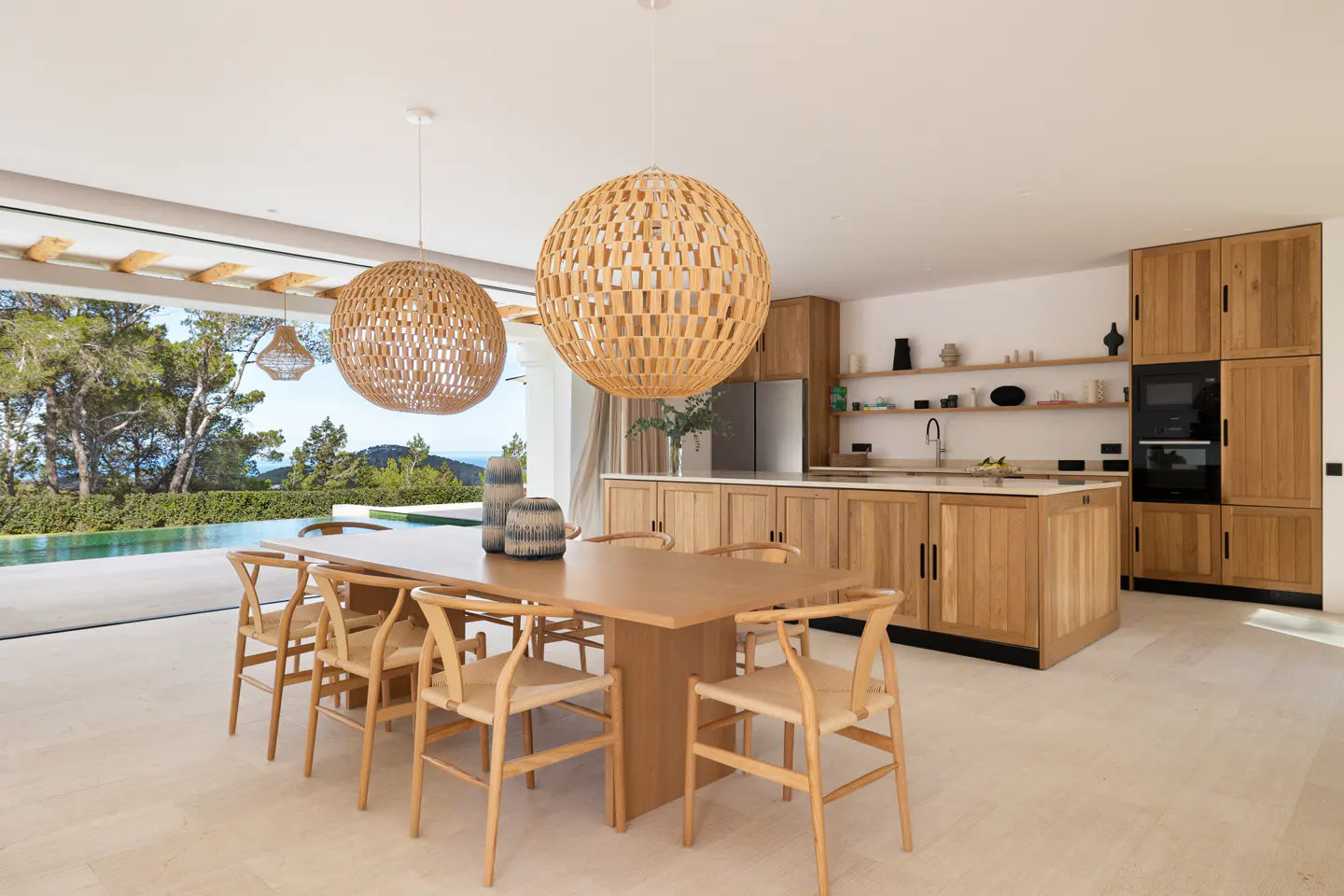 Open-concept kitchen and dining area with wood cabinets, table, and chairs. Large windows overlook a pool and lush greenery. Two woven pendant lights hang above.