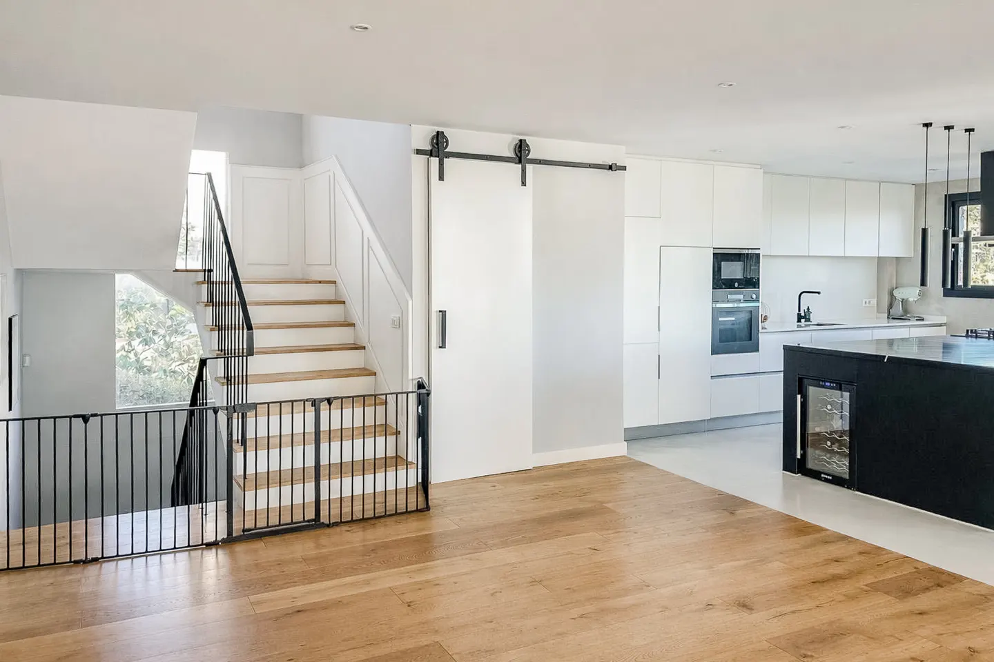Open-concept home interior with wood floors, white walls, and a modern kitchen. Stairs with a black railing lead to the upper level.