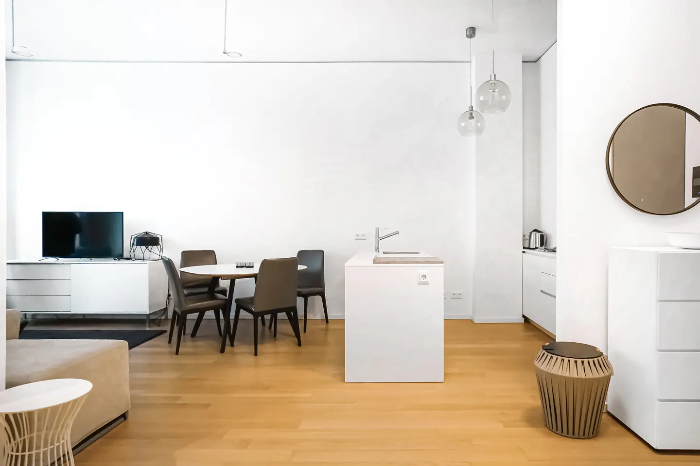 Bright, modern apartment interior with white walls, wood floors, and minimalist furniture. A round dining table and kitchen island are visible.