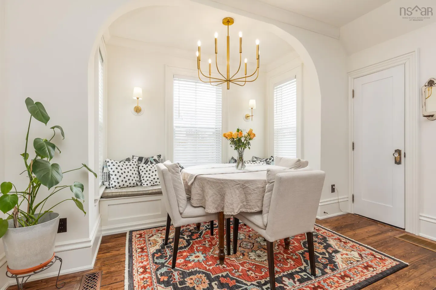Bright dining room with white walls, wood floors, and a colorful rug. A gold chandelier hangs above a round table with four chairs. A window seat is in the background.