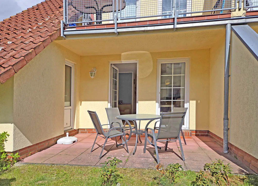 Outdoor patio with a round table and four chairs on a red tiled floor. A yellow building with a red tiled roof is in the background.