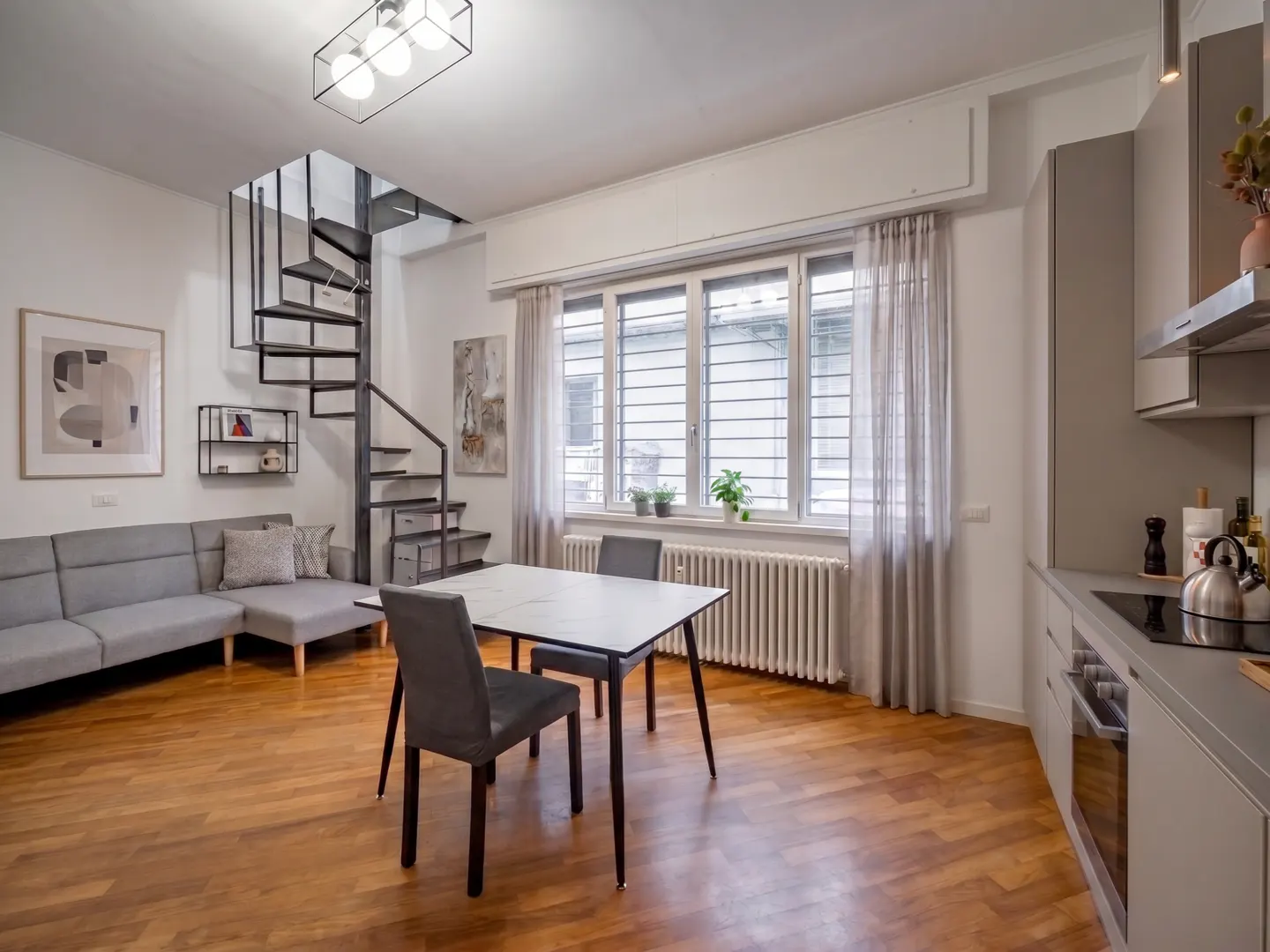 A modern apartment interior with a gray sofa, spiral staircase, dining table, and kitchen area. The room has hardwood floors and natural light from a large window.