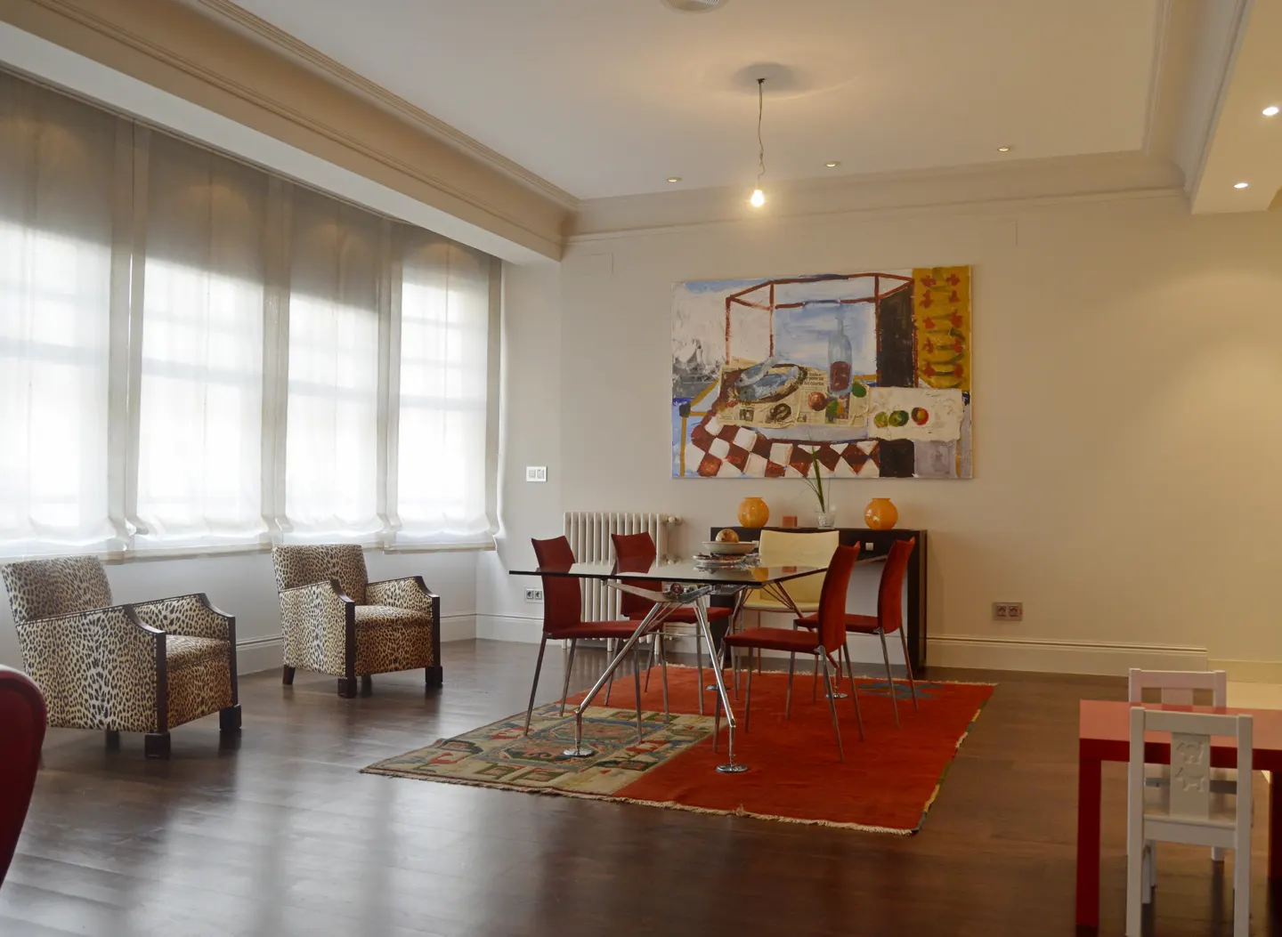 Bright living room with dark wood floors, leopard print chairs, and a glass table with red chairs on a red rug. A large painting hangs on the wall.