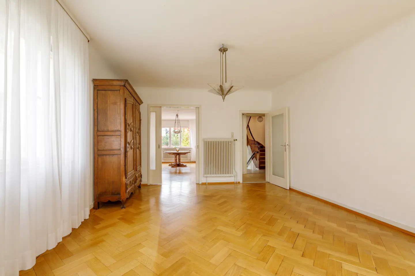 Bright room with herringbone wood floors, white walls, and a large wooden armoire. A doorway leads to a dining room, and another to a staircase.