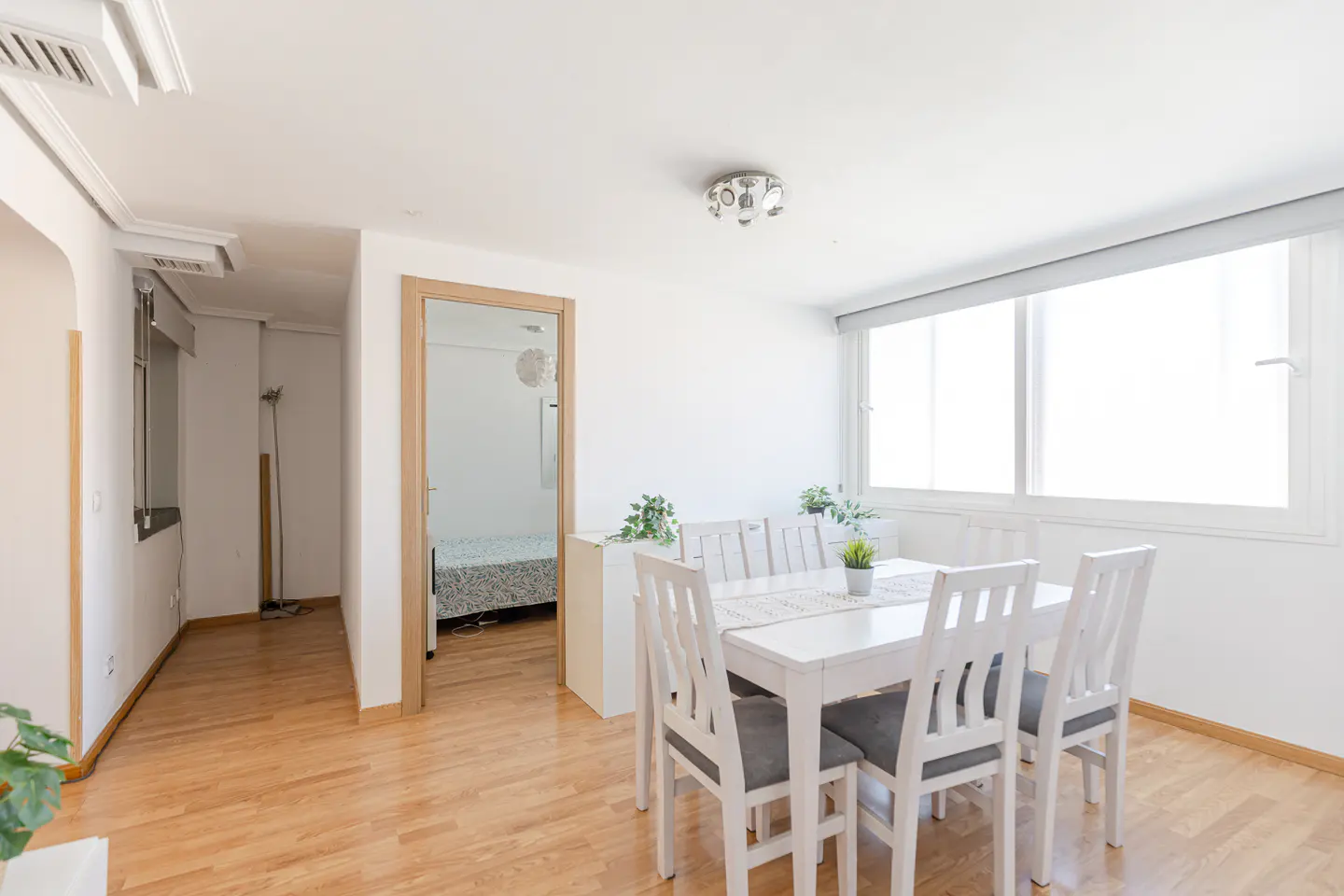Bright dining area with a white table, six chairs with gray cushions, and a large window. A doorway leads to a bedroom. Hardwood floors.
