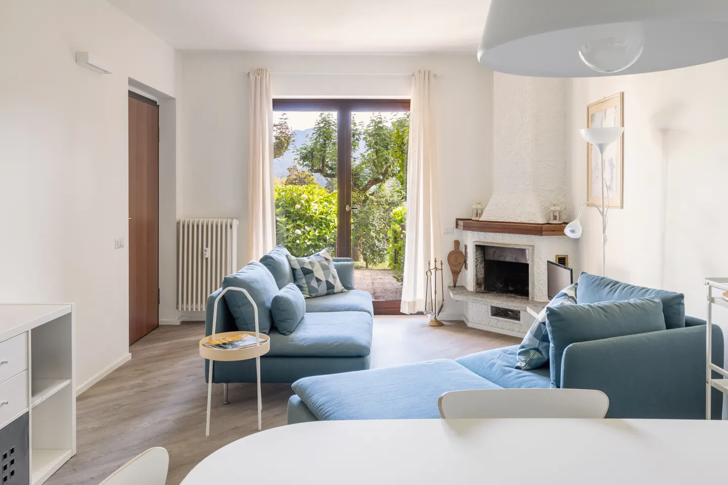Bright living room with a blue sectional sofa, white walls, and a stone fireplace. A glass door leads to a lush green garden.
