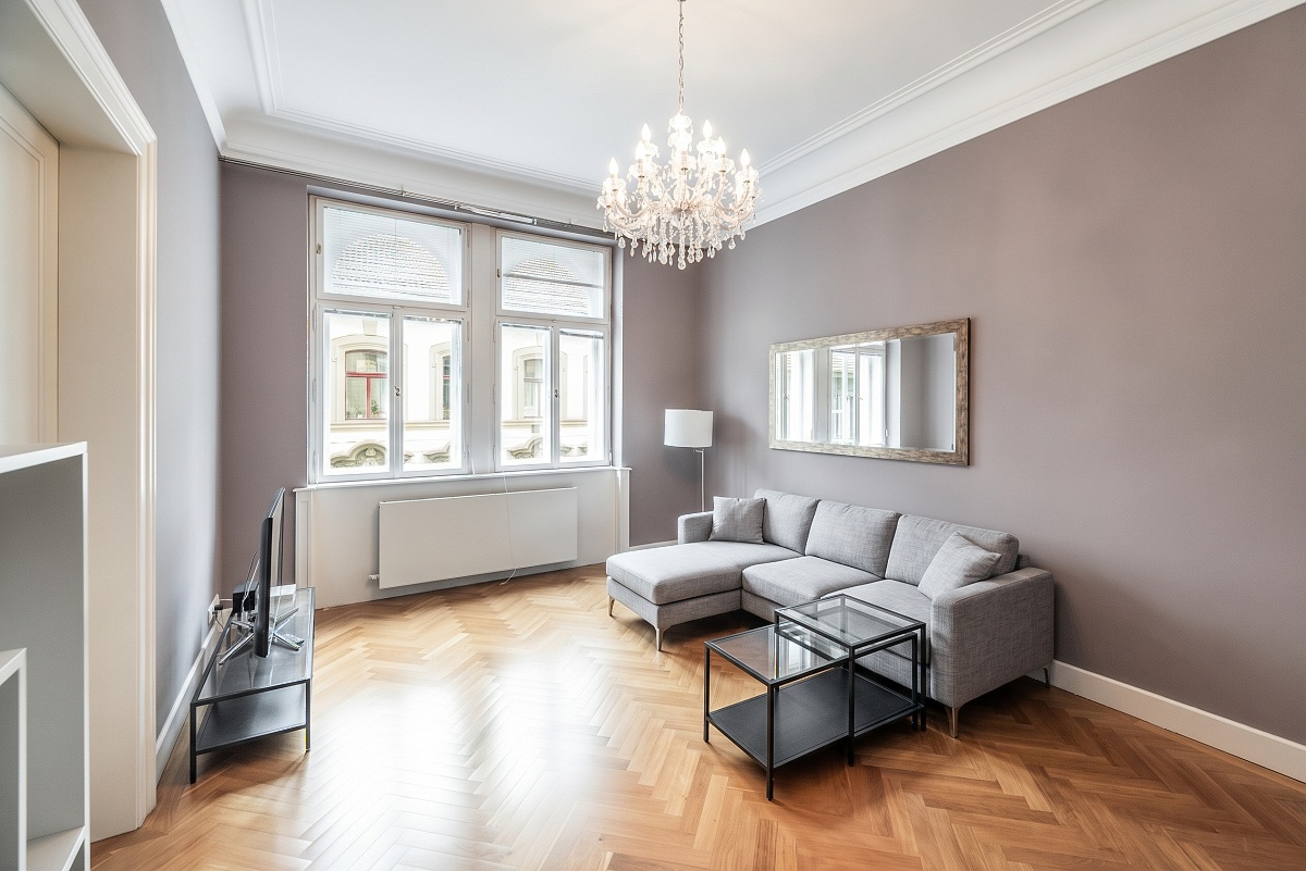 Living room with herringbone wood floors, grey walls, a grey sectional sofa, glass coffee tables, and a crystal chandelier. [AI-generated text]