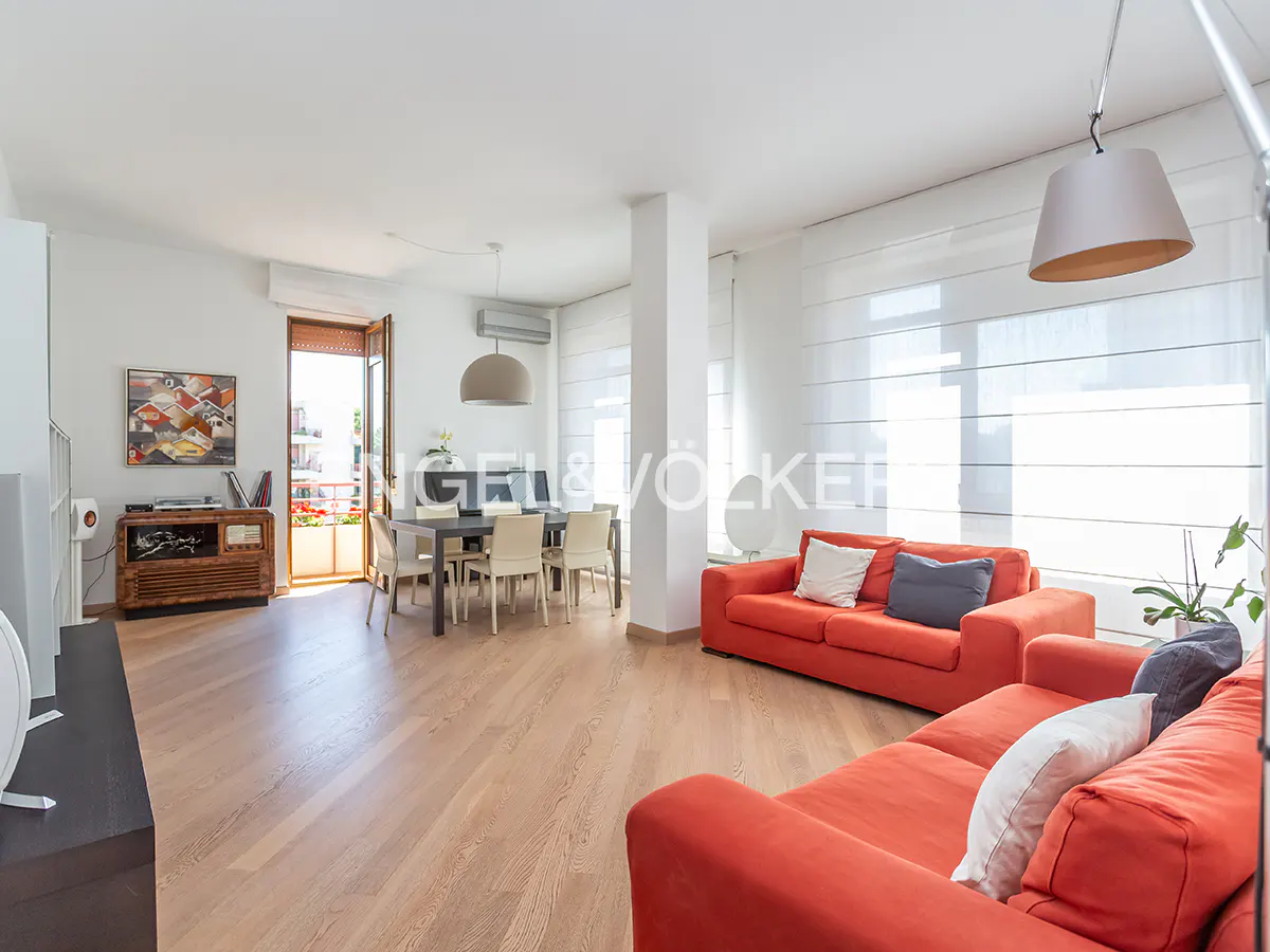 Bright open-plan living room with light wood floors, two orange sofas, a dining area, and a balcony door.