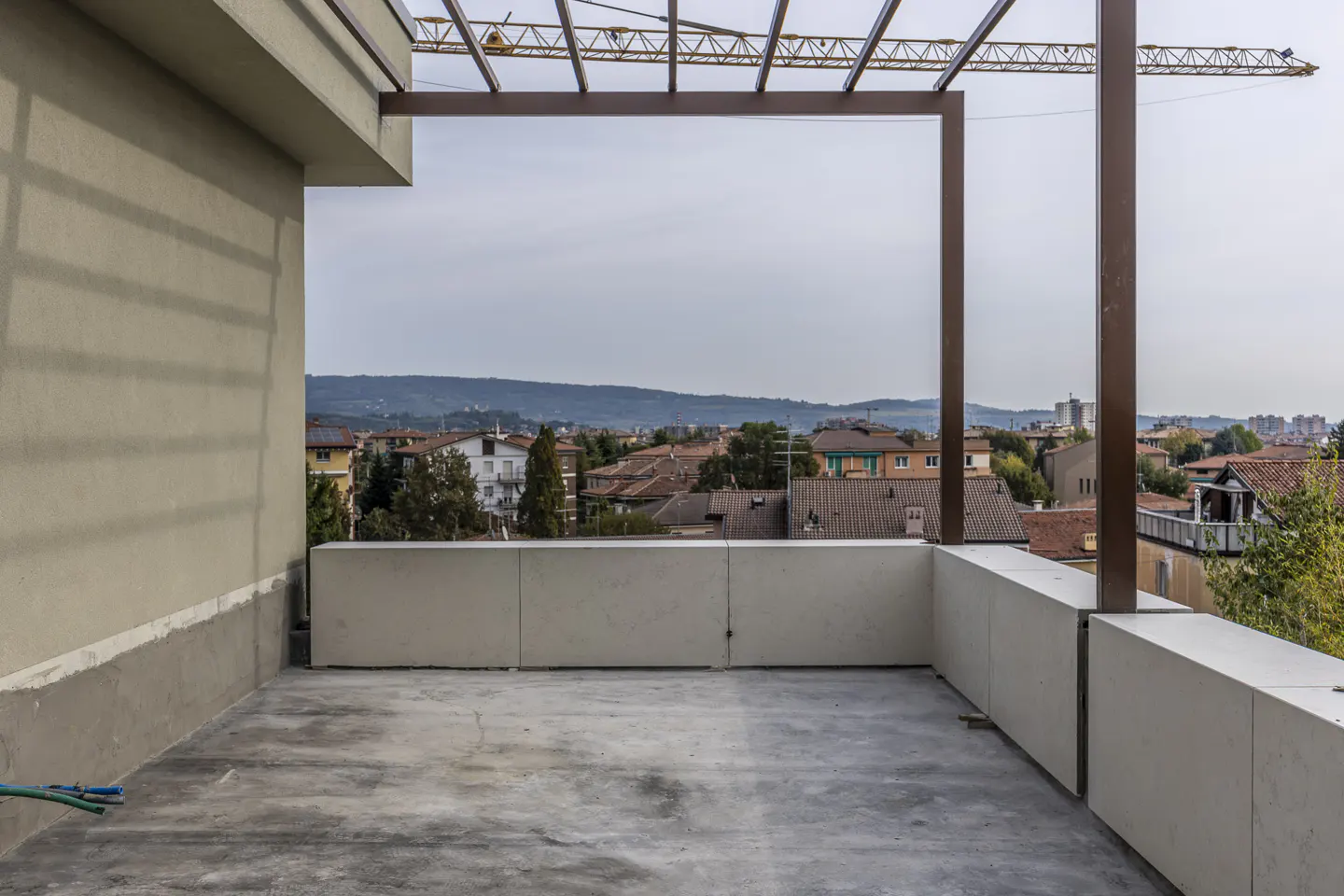 Unfinished balcony with concrete floor and low wall overlooking a cityscape with red-tiled roofs and distant hills under a cloudy sky.