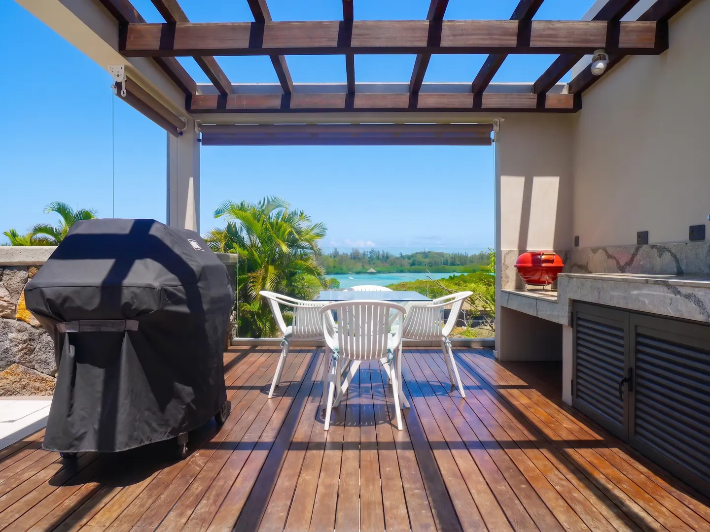 Outdoor patio with wooden pergola, grill, and dining set overlooking a blue ocean and lush greenery.
