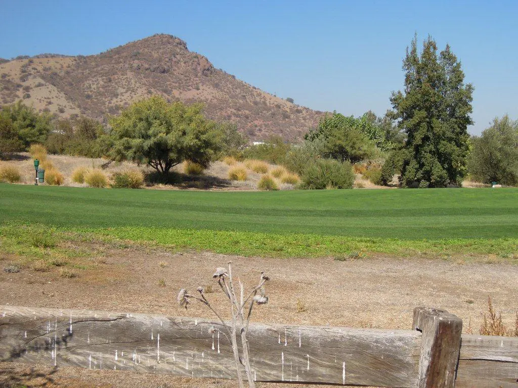 A green golf course with a mountain backdrop under a blue sky. A weathered wooden fence is in the foreground.