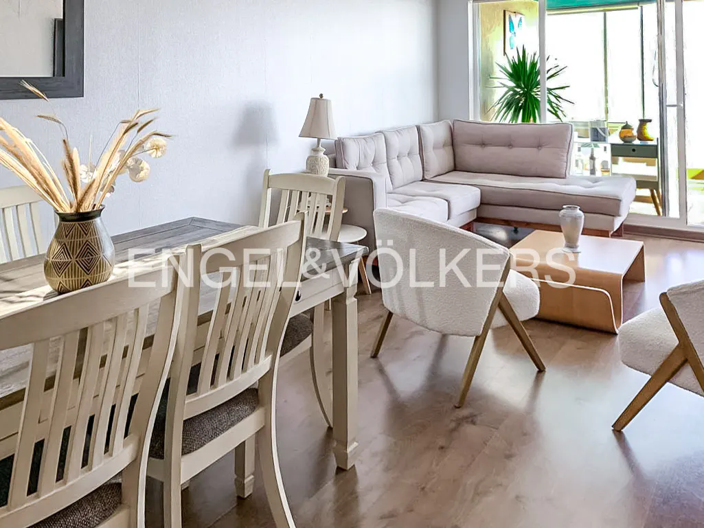 Living room with dining table, beige sectional sofa, white chairs, wood floors, and a balcony with plants.