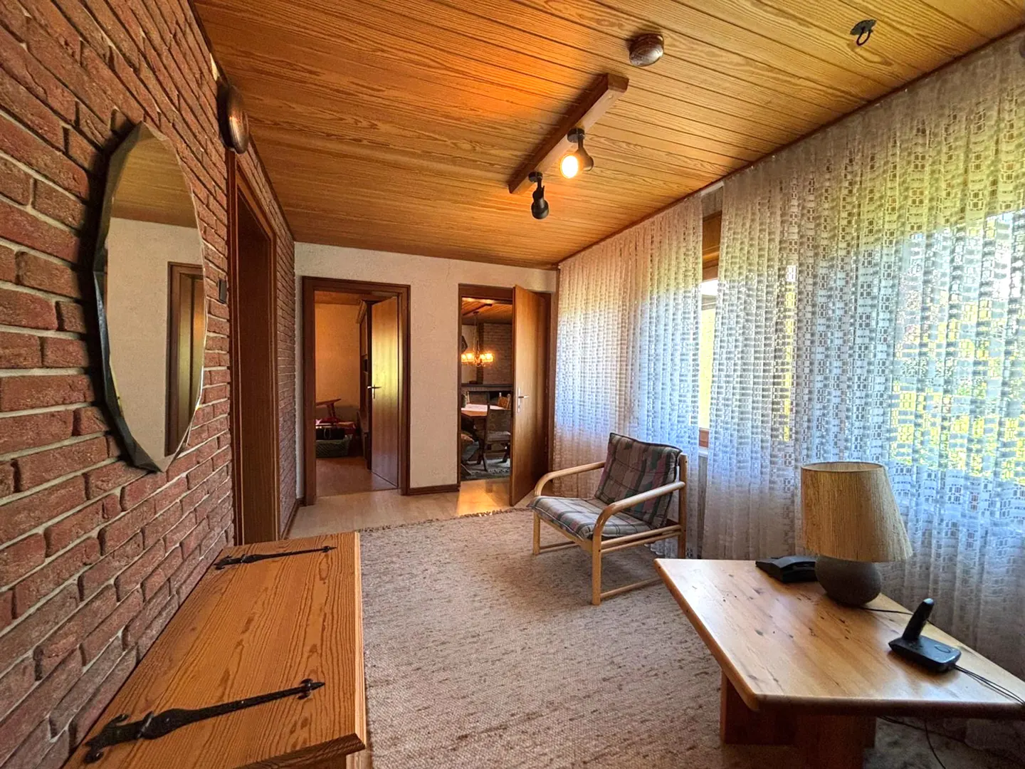 A cozy room with a brick wall, wood ceiling, and woven curtains. A wooden bench, chair, and table furnish the space.