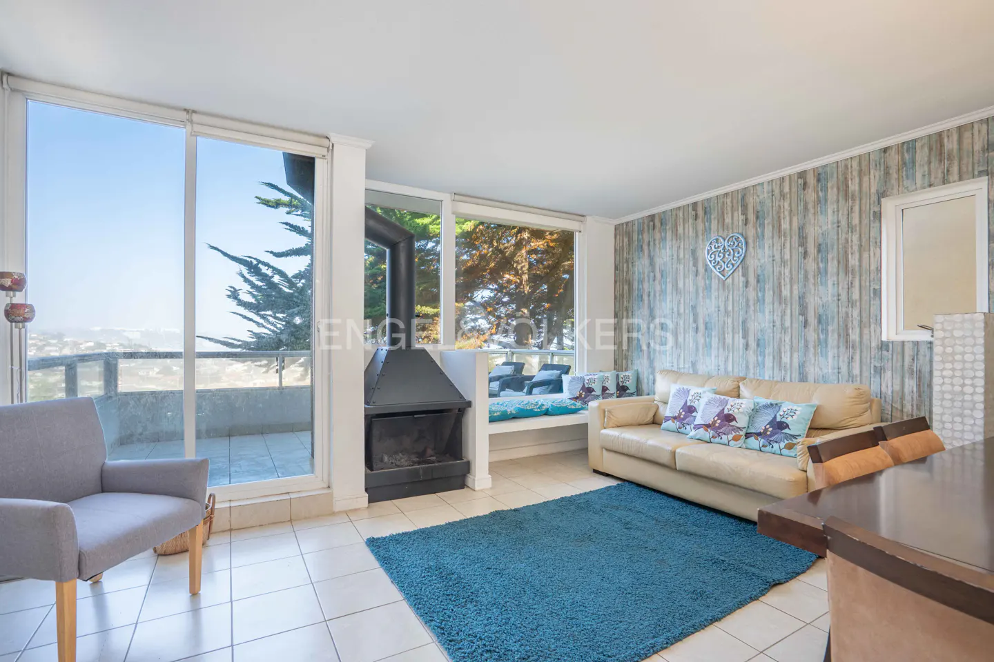 Living room with beige sofa, blue rug, grey armchair, and a modern fireplace, set against a wall with ocean-view windows.