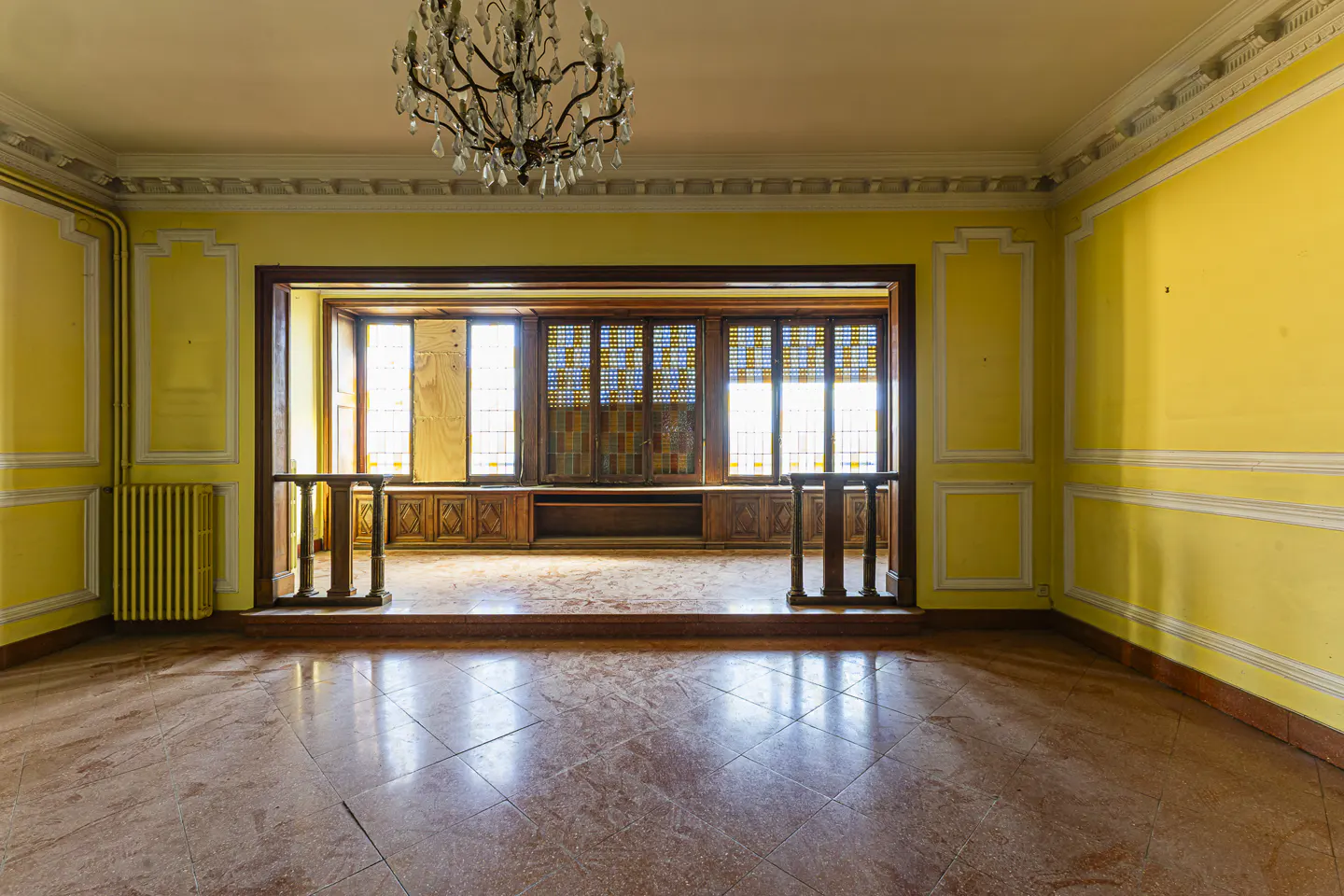 A yellow room with a chandelier and a window seat. The floor is tiled in a pink marble pattern.
