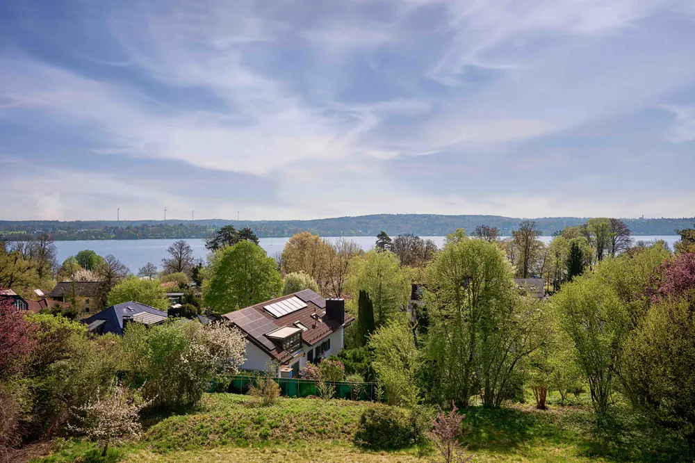 Scenic view of a house with solar panels, surrounded by lush trees, overlooking a lake under a partly cloudy sky.