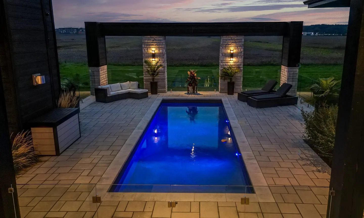 Outdoor pool with blue lighting, stone pillars, and lounge furniture on a paved patio overlooking a green field at dusk.