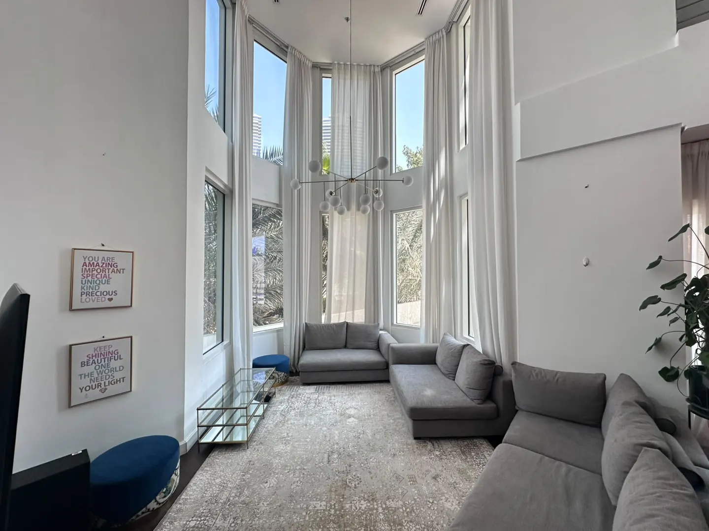 Modern living room with high ceilings, floor-to-ceiling windows, grey sectional sofas, and a contemporary chandelier.
