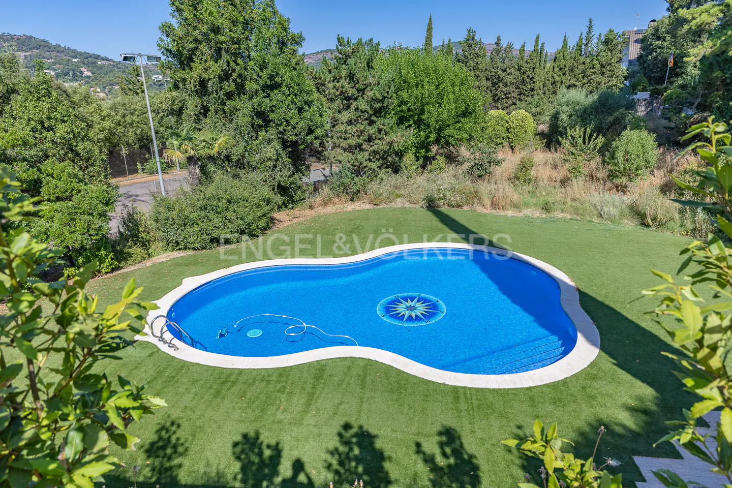 Aerial view of a blue kidney-shaped pool with a star mosaic, surrounded by green grass and trees. Engel & Völkers logo visible.