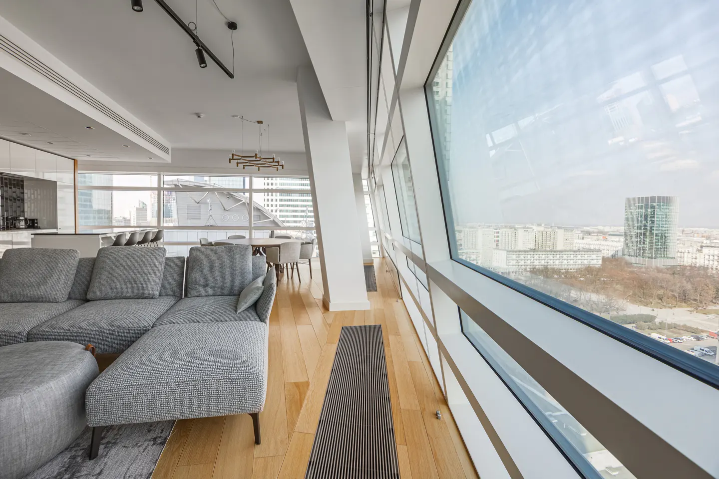 Bright, modern apartment interior with a gray sectional sofa, wood floors, and floor-to-ceiling windows overlooking a city skyline.