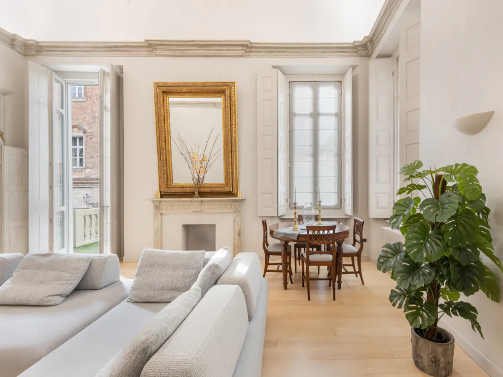Bright living room with a gray sofa, wood floors, and white walls. A gold-framed mirror hangs above a fireplace. A round table with chairs sits near a window. A large potted plant adds greenery.