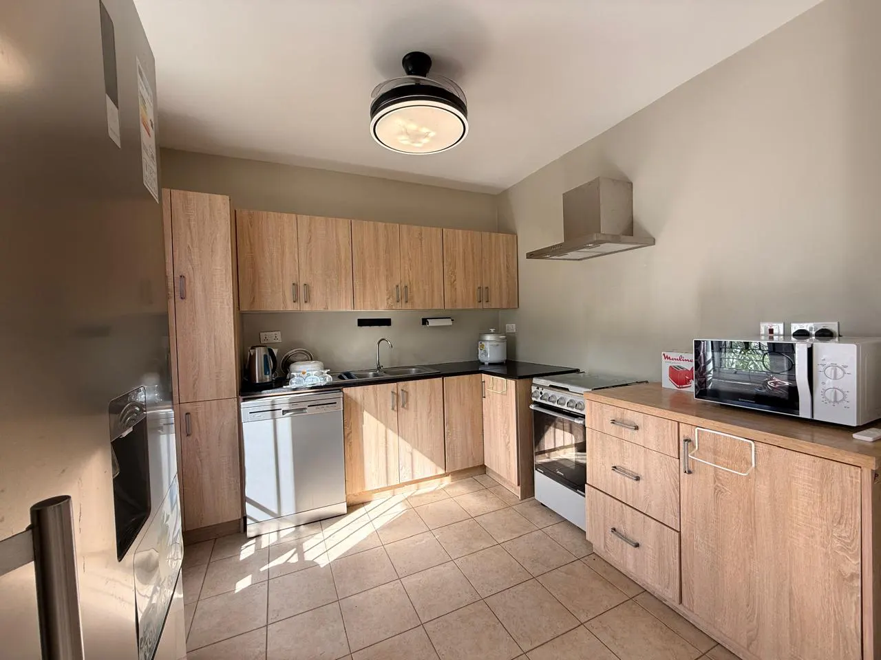 A well-lit kitchen with light wood cabinets, stainless steel appliances, and beige tile flooring.