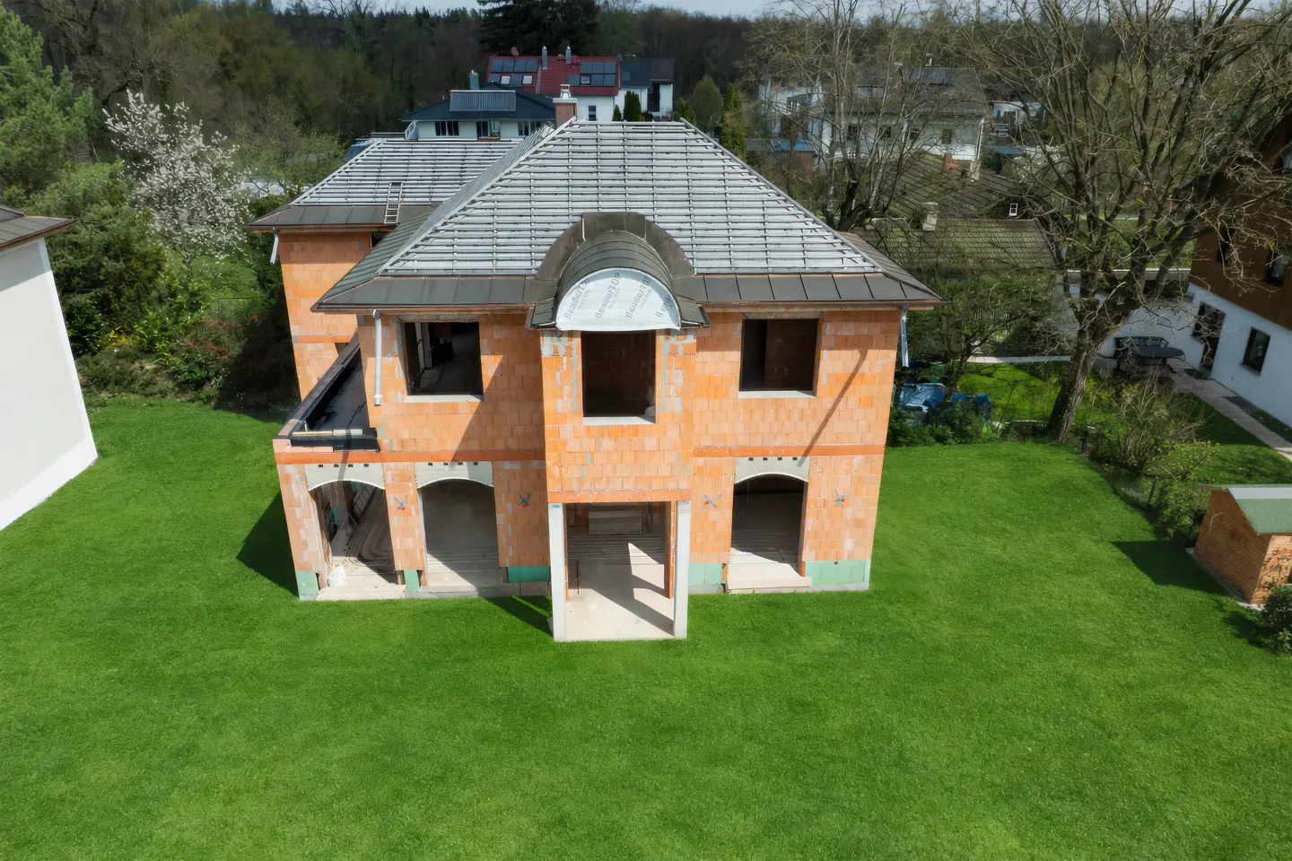 Aerial view of a two-story orange brick house under construction with a gray roof, surrounded by a green lawn and trees.