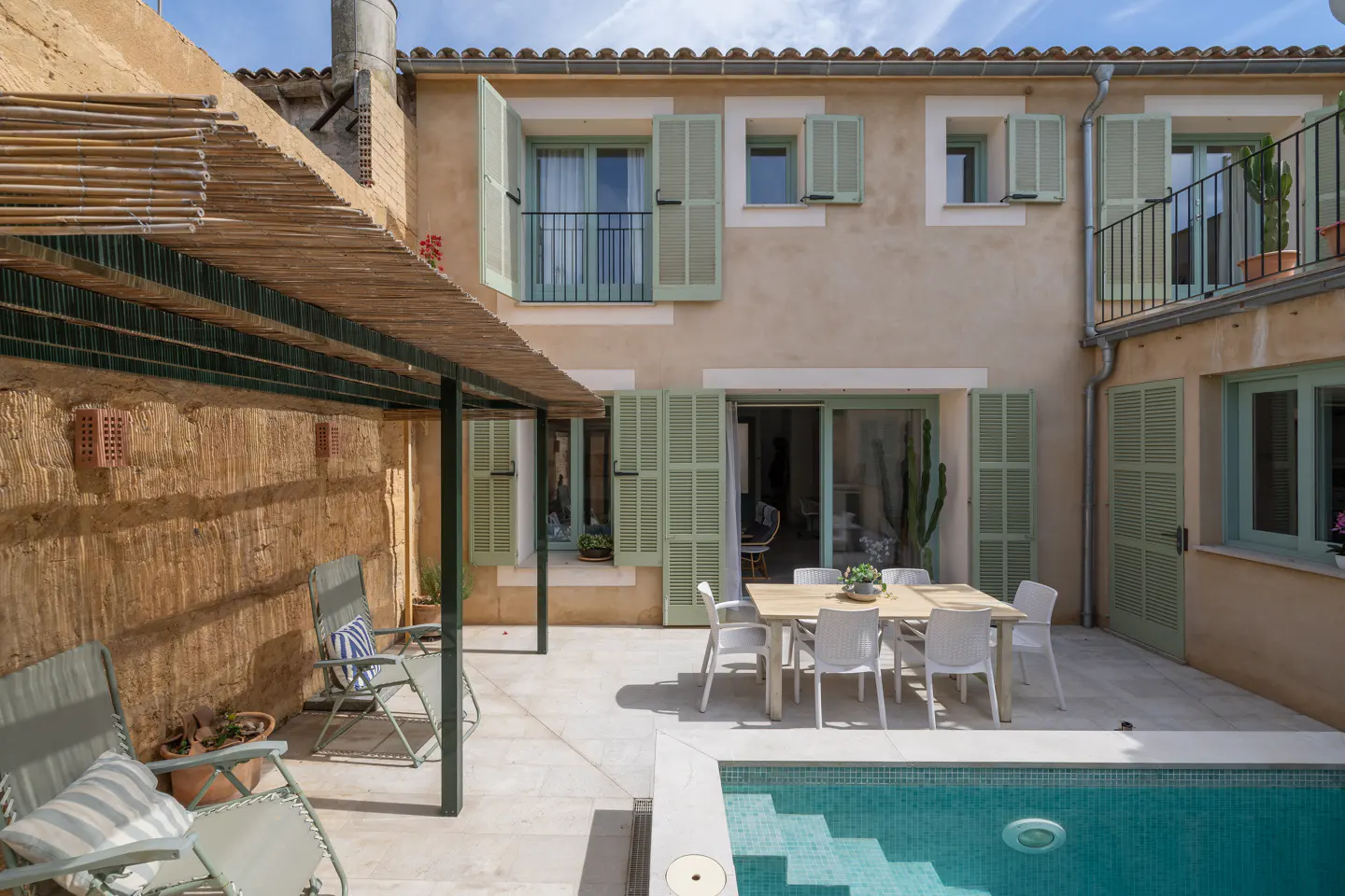 Outdoor patio with a pool, dining table, and lounge chairs. The building has light green shutters and a balcony.