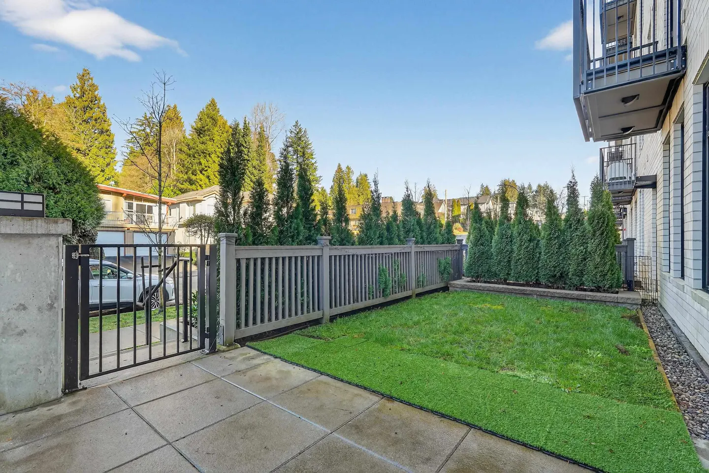 A fenced backyard with green grass, trees, and a patio. A black metal gate leads to the yard. Balconies are visible on the building.
