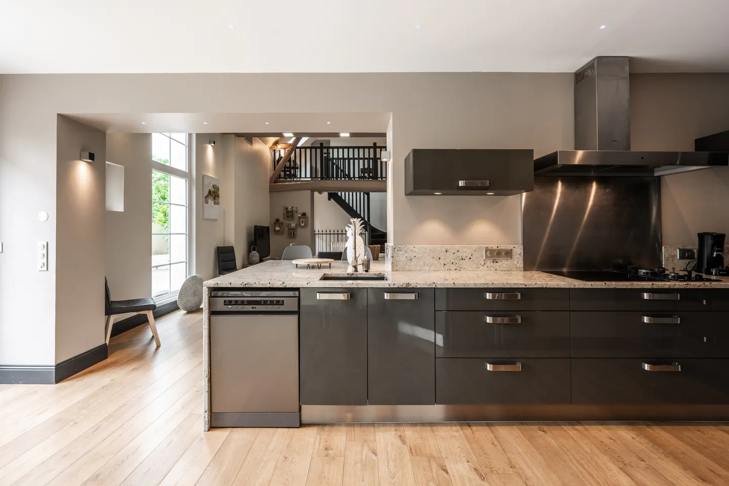 A modern kitchen with gray cabinets, speckled countertops, and stainless steel appliances. A wooden floor and a view to a second-floor loft.