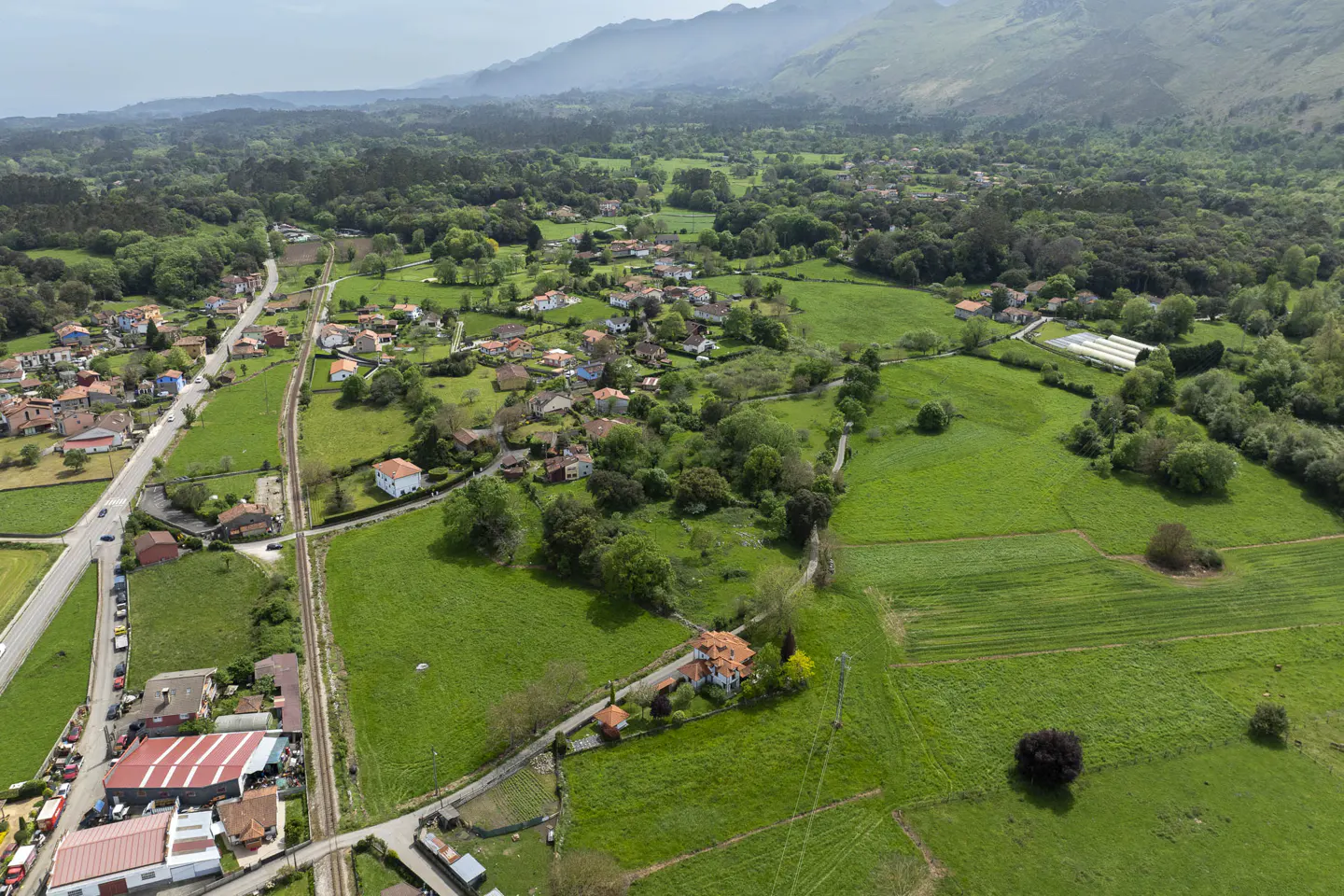 Aerial view of a rural landscape with green fields, trees, houses with red roofs, a road, and mountains in the background.
