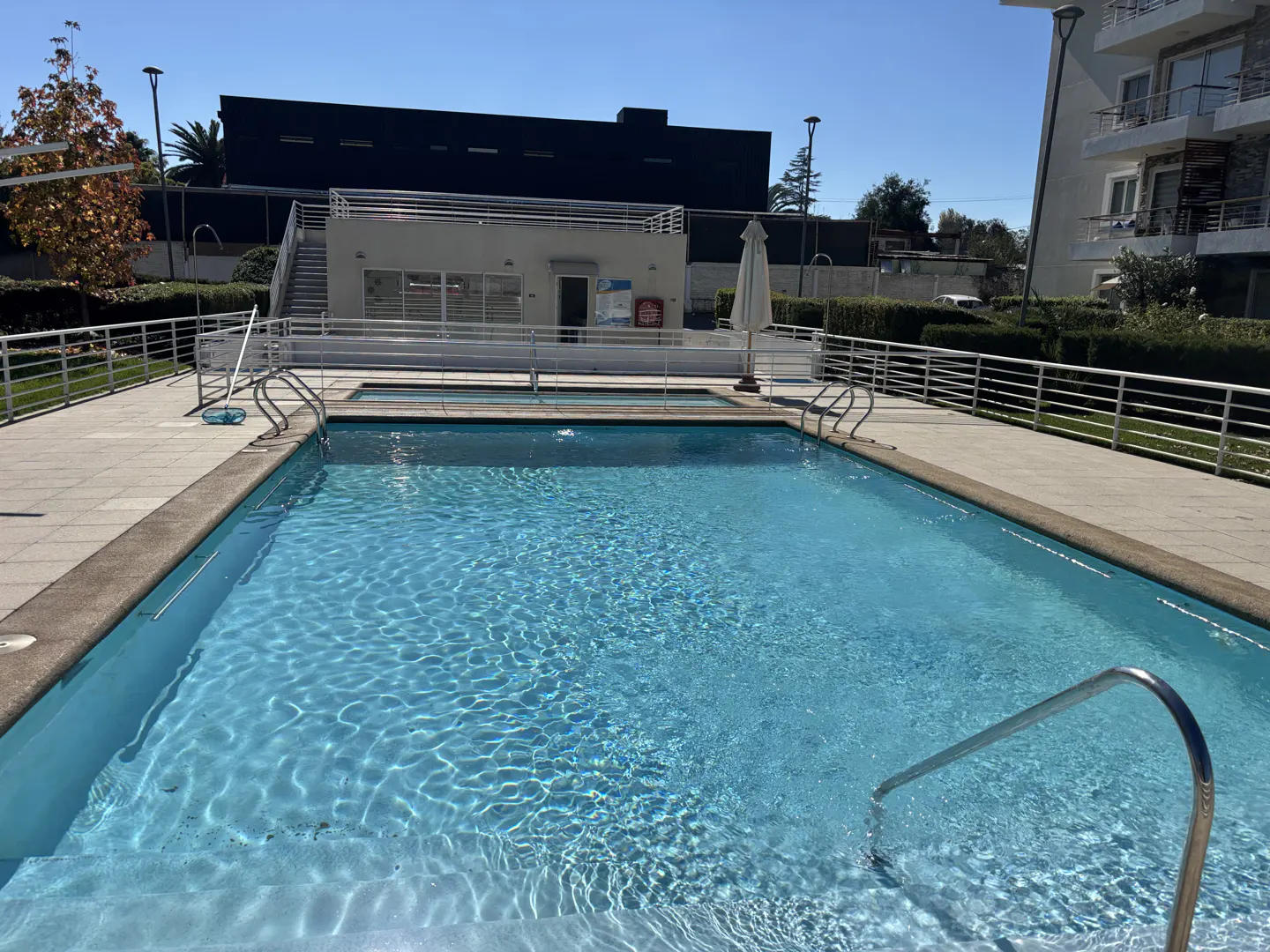 Outdoor pool with clear blue water, metal railings, and a pool house in the background on a sunny day.