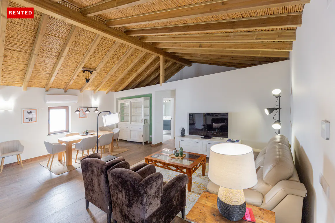 A bright living and dining area with a wooden ceiling, brown chairs, and a beige sofa. A "RENTED" tag is in the upper left corner.