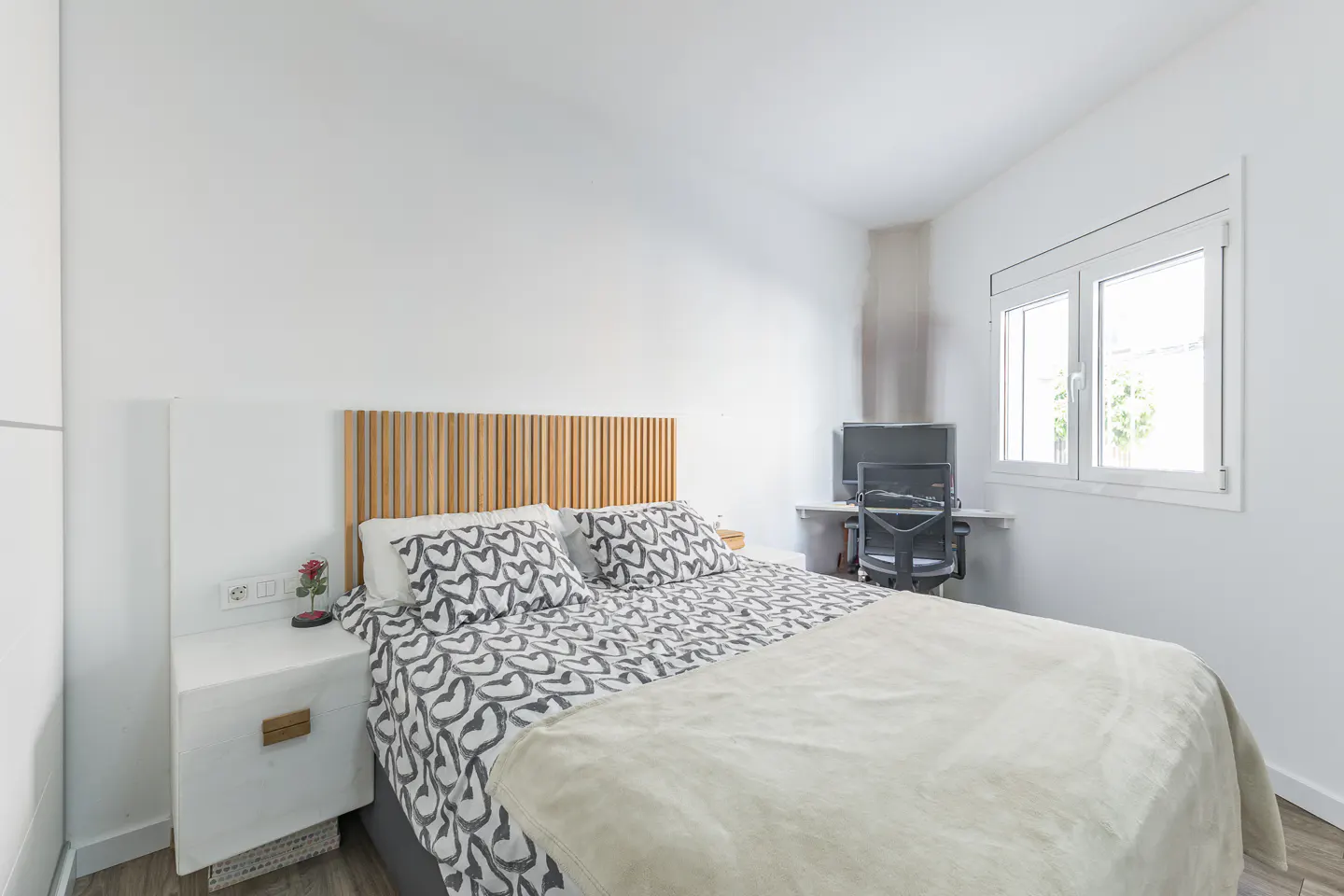 Bright bedroom with a bed featuring a wooden headboard and heart-patterned bedding, plus a desk with a computer by a window.