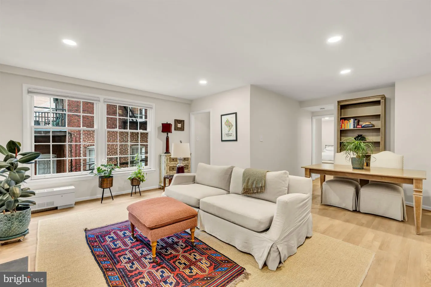 Bright living room with a beige sofa, ottoman, and patterned rug. Large windows offer a view of a brick building. A wooden table and bookshelf are in the background.