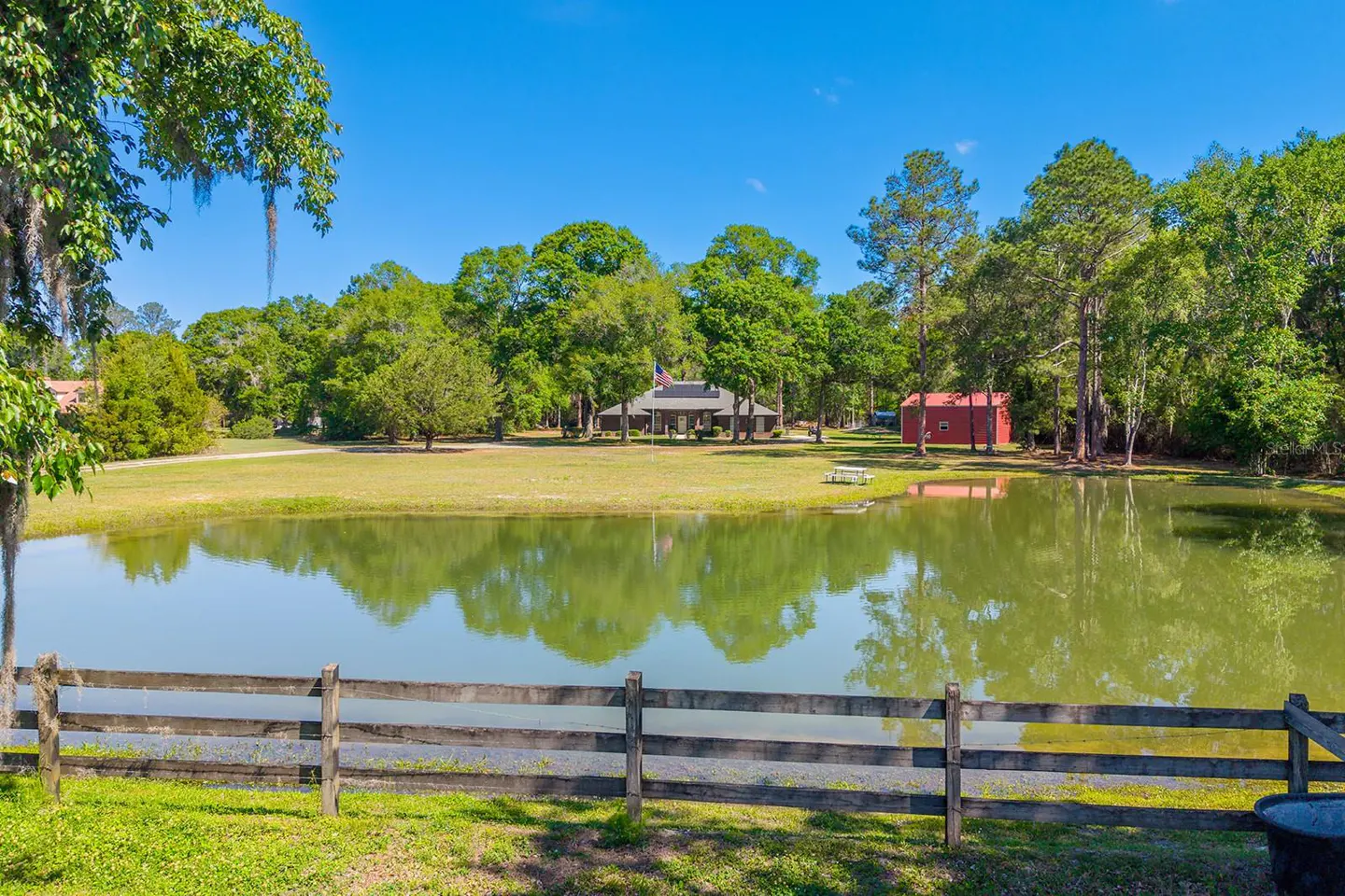 Scenic view of a pond reflecting trees and a house, framed by a wooden fence in the foreground.