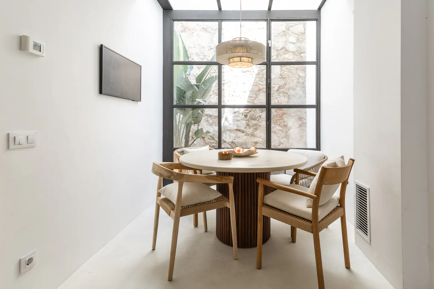 Bright dining area with a round table, four chairs, and a large window with a view of greenery and a stone wall. A modern light fixture hangs above.