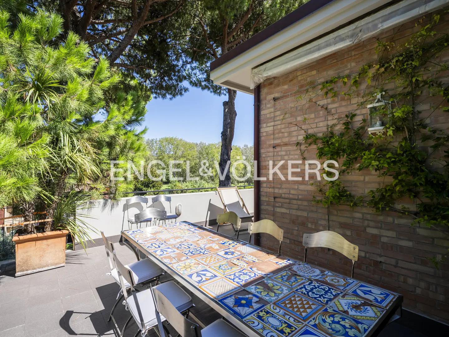 Outdoor patio with a long table covered in colorful tiles, surrounded by chairs. Brick wall with vines and trees in the background.