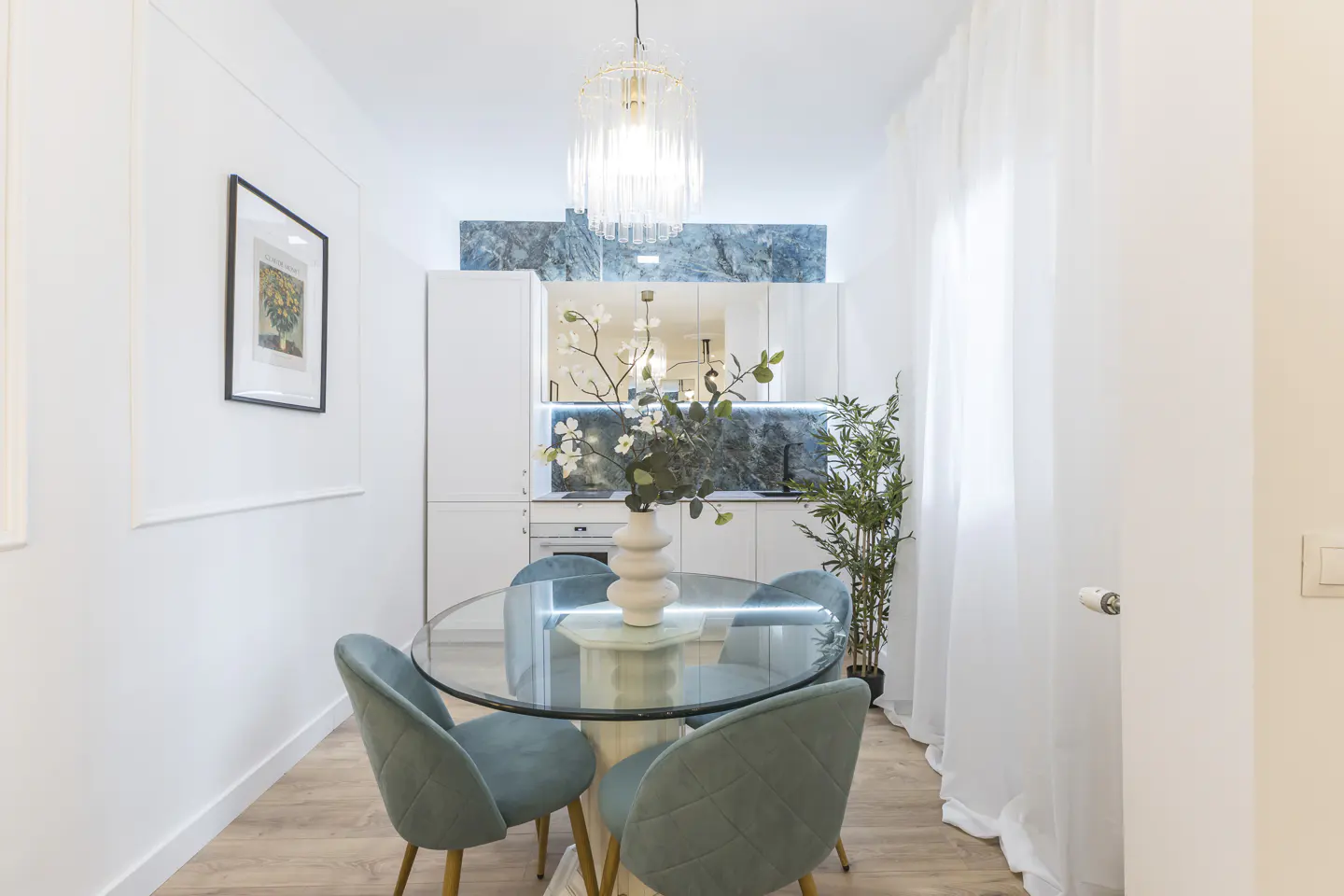 Bright dining area with a glass table, four teal chairs, and a white vase with flowers. A modern chandelier hangs above, with white cabinets and marble backsplash in the background.