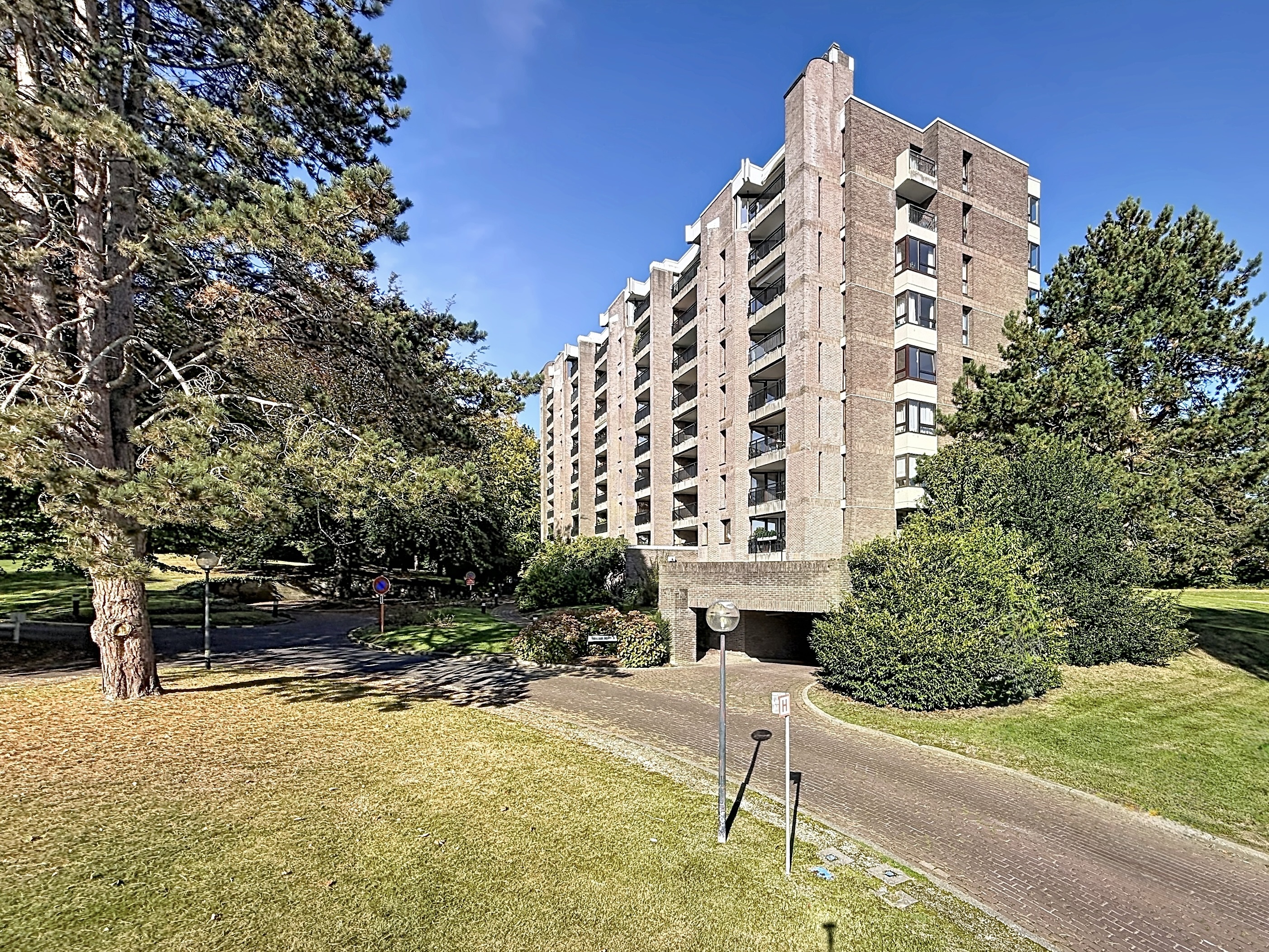 Tall brown brick apartment building with balconies, surrounded by green trees and a paved driveway under a clear blue sky. [AI-generated text]