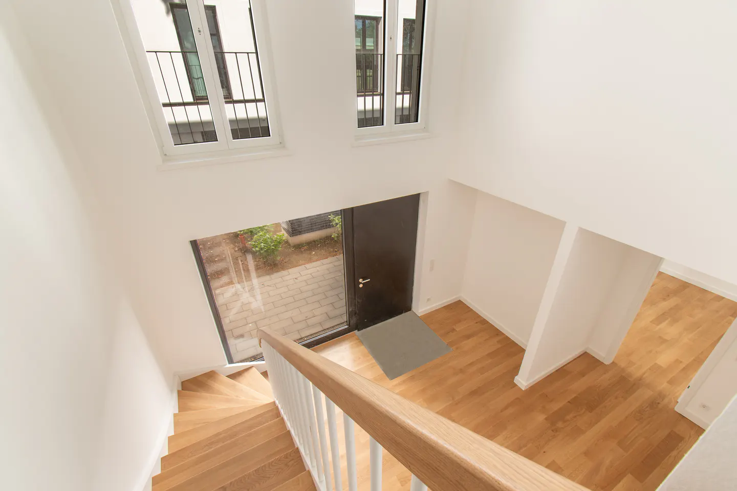 A high-angle shot of a modern home interior with wooden stairs, floors, and white walls. Windows and a black door are visible.