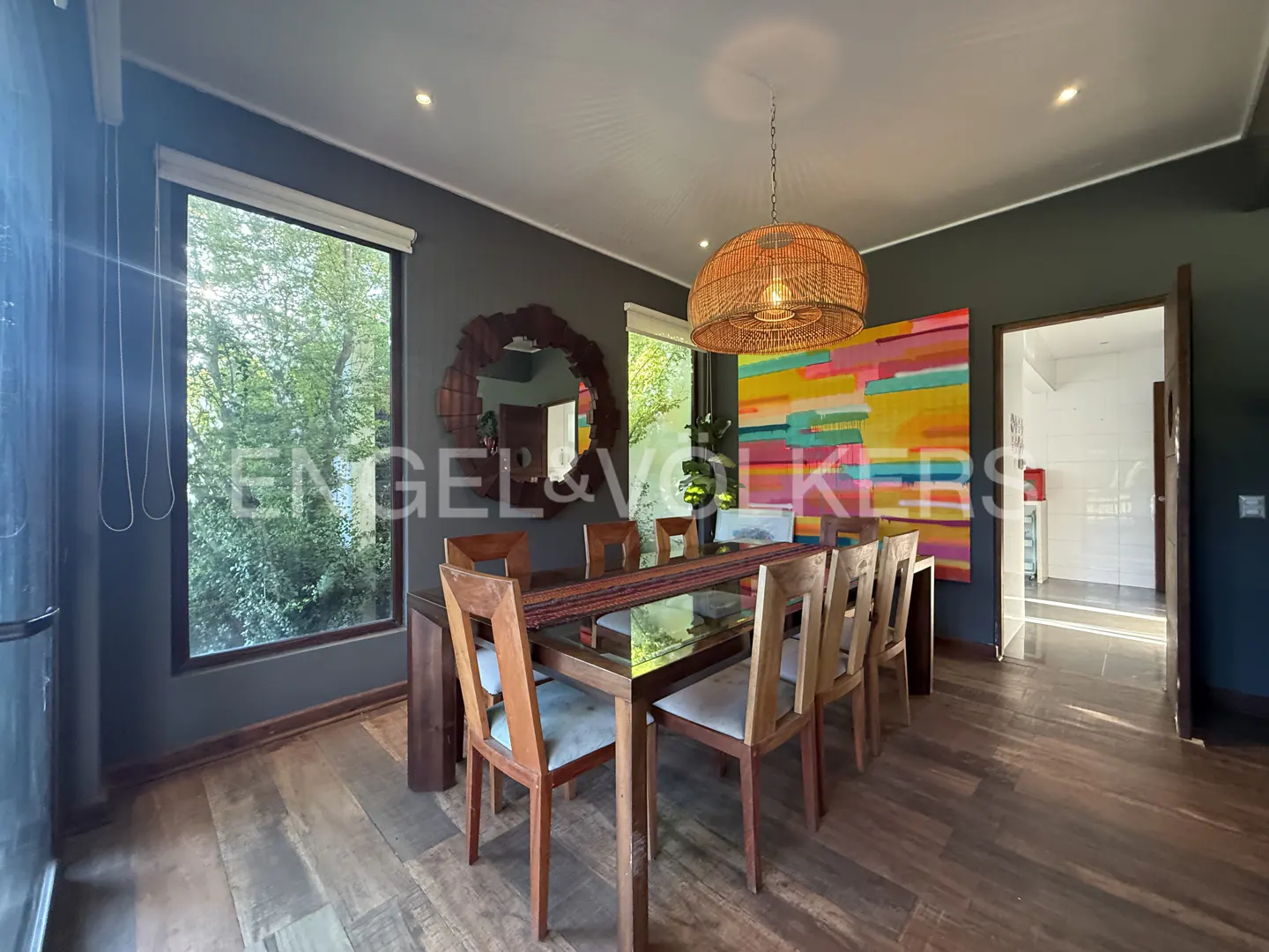 Dining room with wood table, chairs, and floor. A woven pendant light hangs above. A colorful painting is on the wall.