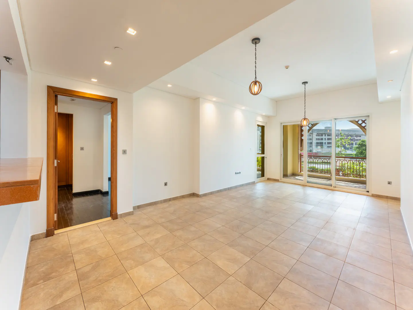 Bright, empty living room with beige tile floor, white walls, and balcony access. Two pendant lights hang from the ceiling.