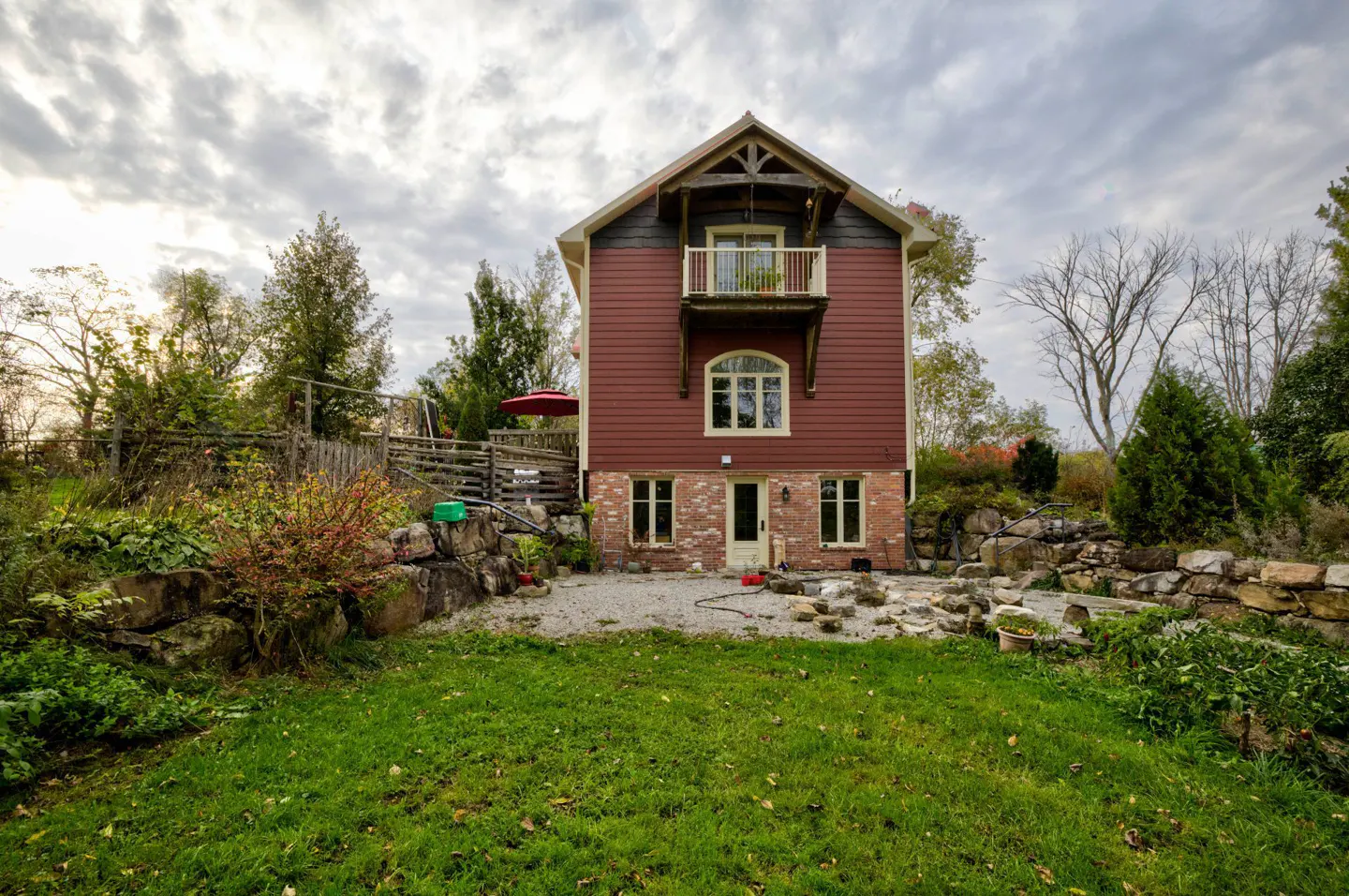 Two-story house with red siding, brick base, and a small balcony, surrounded by a green lawn and rock gardens.