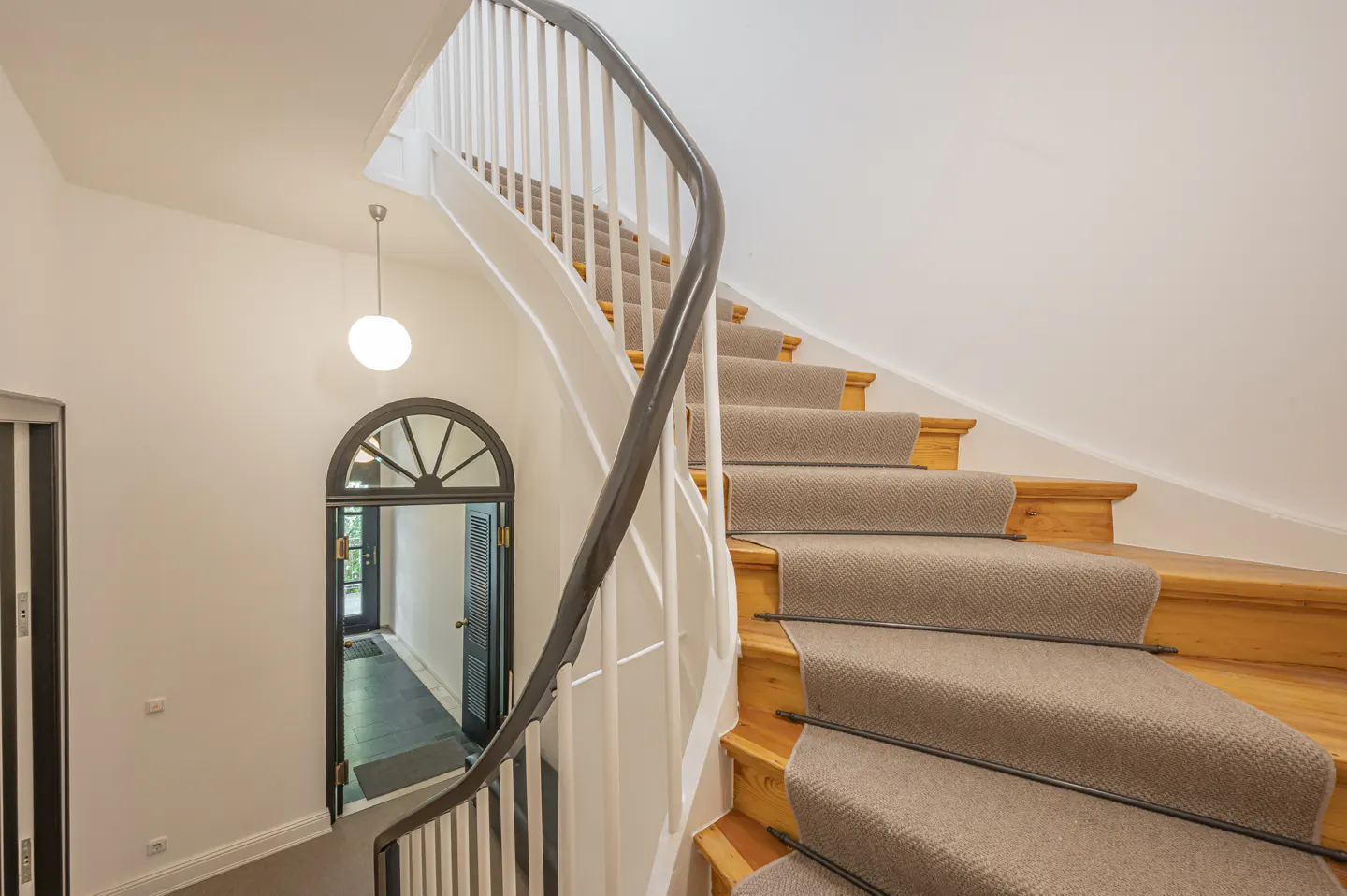 Interior view of a curved staircase with a beige runner, white walls, and a dark wood railing. A doorway with an arched window is visible.