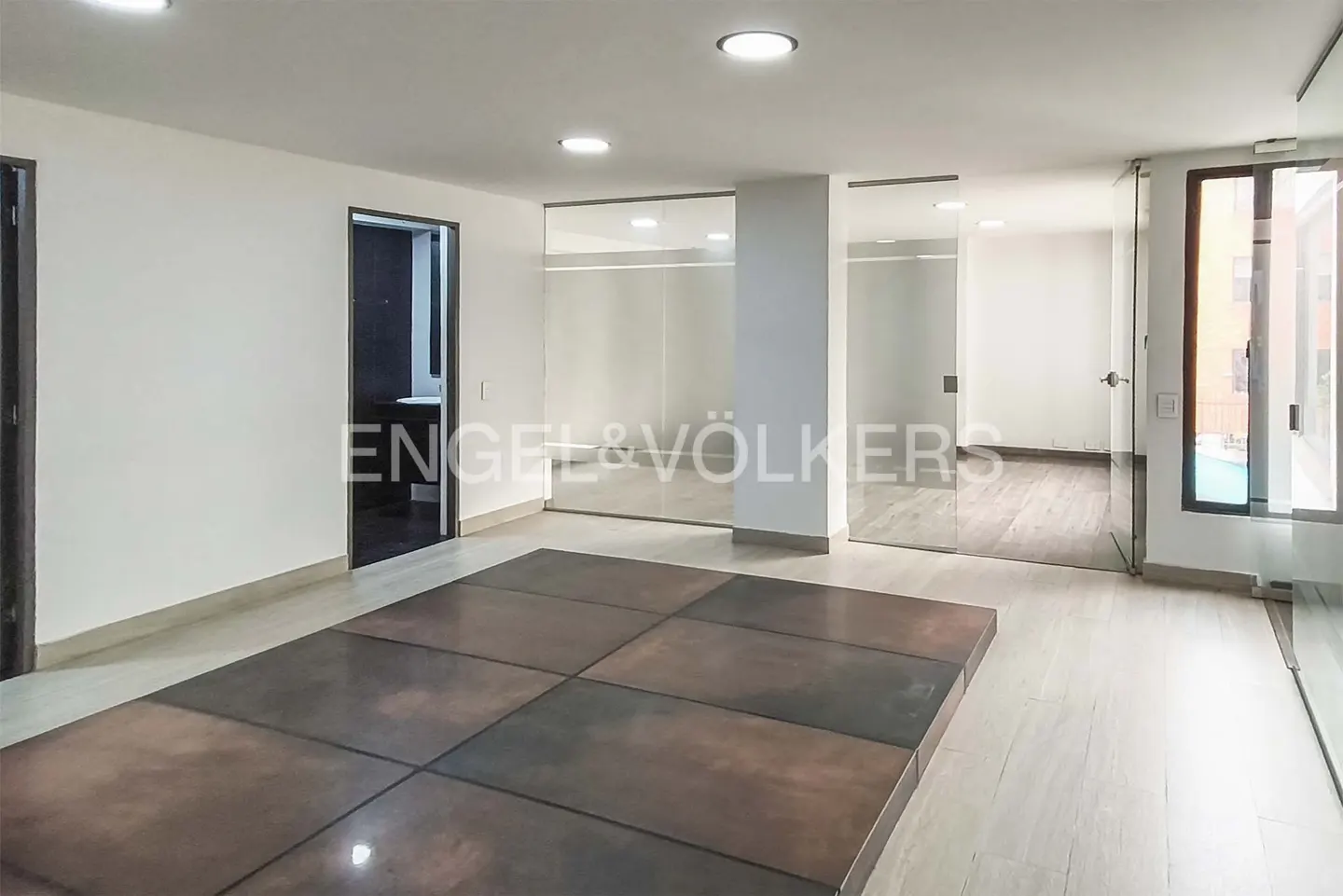 An empty room with white walls, glass doors, and a brown tiled floor. The Engel & Völkers logo is visible in the center.